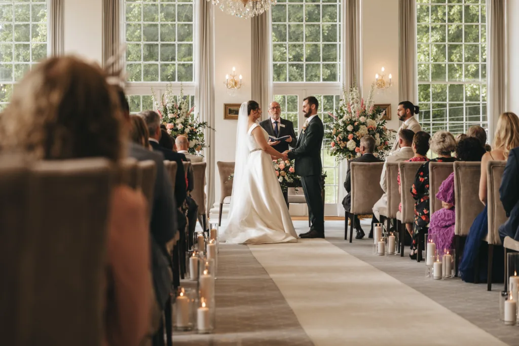 A bride and groom stand facing each other at the altar in a bright, elegant room with tall windows. Guests are seated on both sides of a candle-lined aisle as an east midlands wedding photographer captures candid moments of the beautiful ceremony. © Aimee Lince Photography - Wedding photographer in Lincolnshire, Yorkshire & Nottinghamshire