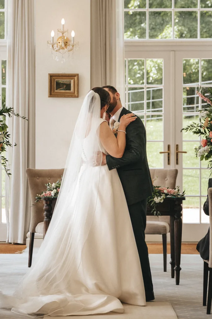 A bride in a white gown and veil kisses a groom in a dark suit during an indoor wedding ceremony. Captured by an East Midlands wedding photographer, the candid wedding photography highlights their love amid classic decor and soft natural light. © Aimee Lince Photography - Wedding photographer in Lincolnshire, Yorkshire & Nottinghamshire