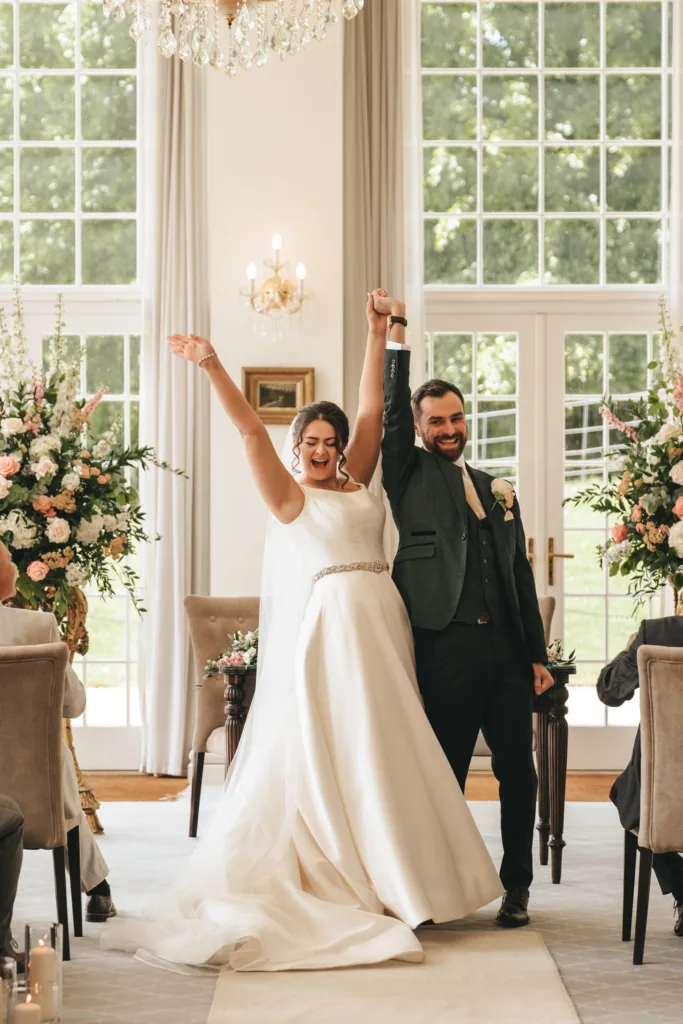 A bride in a white gown and a groom in a dark suit stand at the aisle, joyfully raising clasped hands. Captured by a candid wedding photography expert, they celebrate among lush florals and guests as sunlight streams through tall windows. © Aimee Lince Photography - Wedding photographer in Lincolnshire, Yorkshire & Nottinghamshire