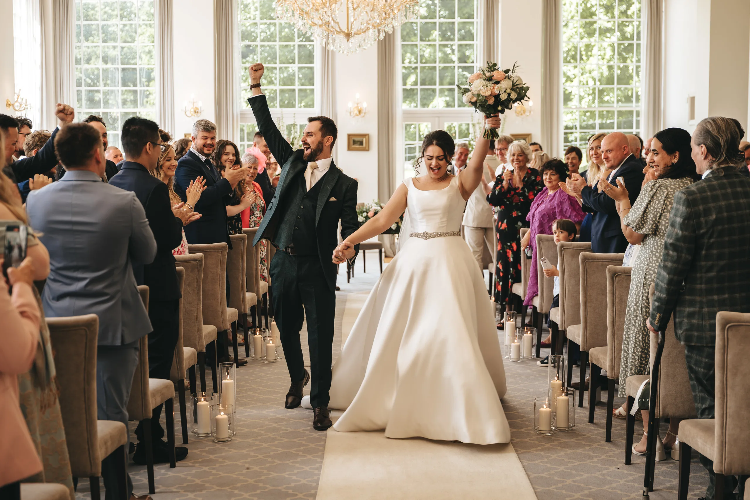 katie-adair-wedding-photography-rushton-hall-kettering-northamptonshire-276-of-1141-©-aimee-lince-photography-02-06-2025 A joyful bride and groom walk down the aisle, holding hands and raising their arms in celebration, surrounded by applauding guests. This candid wedding photography captures pure happiness in a bright, elegant room with large windows and chandeliers. © Aimee Lince Photography - Wedding photographer in Lincolnshire, Yorkshire & Nottinghamshire