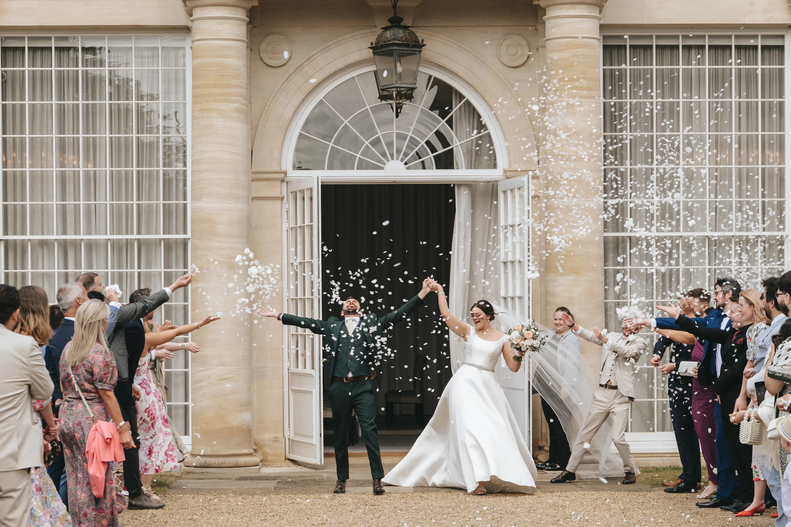katie-adair-wedding-photography-rushton-hall-kettering-northamptonshire-282-of-1141-©-aimee-lince-photography-04-06-2025 A bride and groom joyfully exit Rushton Hall with guests on both sides throwing white confetti. The wedding photographer captures the moment as the couple celebrates outside the grand venue, surrounded by love and happiness. © Aimee Lince Photography - Wedding photographer in Lincolnshire, Yorkshire & Nottinghamshire