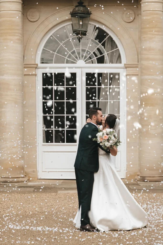 A bride and groom kiss outside elegant Rushton Hall in Kettering, framed by large arched windows. The groom wears a dark suit; the bride holds a bouquet as white confetti falls, captured beautifully by their photographer. © Aimee Lince Photography - Wedding photographer in Lincolnshire, Yorkshire & Nottinghamshire