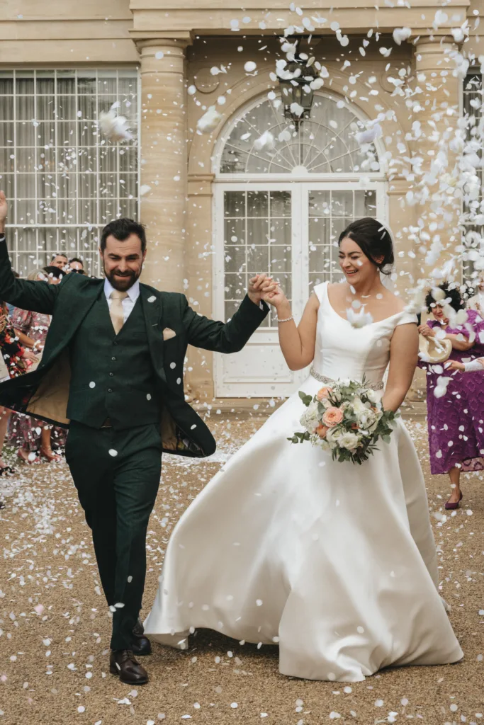 A joyful bride and groom walk hand in hand outside Rushton Hall, smiling as guests shower them with white flower petals. The bride wears an elegant white gown; the groom is in a dark suit—the perfect moment for any wedding photographer. © Aimee Lince Photography - Wedding photographer in Lincolnshire, Yorkshire & Nottinghamshire