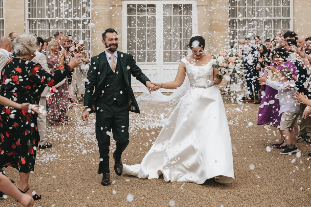 A bride in a white satin gown and a groom in a dark suit hold hands and smile as guests throw white confetti outside a grand building in Kettering. The wedding photographer captures guests clapping and celebrating by the large windows and double doors. © Aimee Lince Photography - Wedding photographer in Lincolnshire, Yorkshire & Nottinghamshire