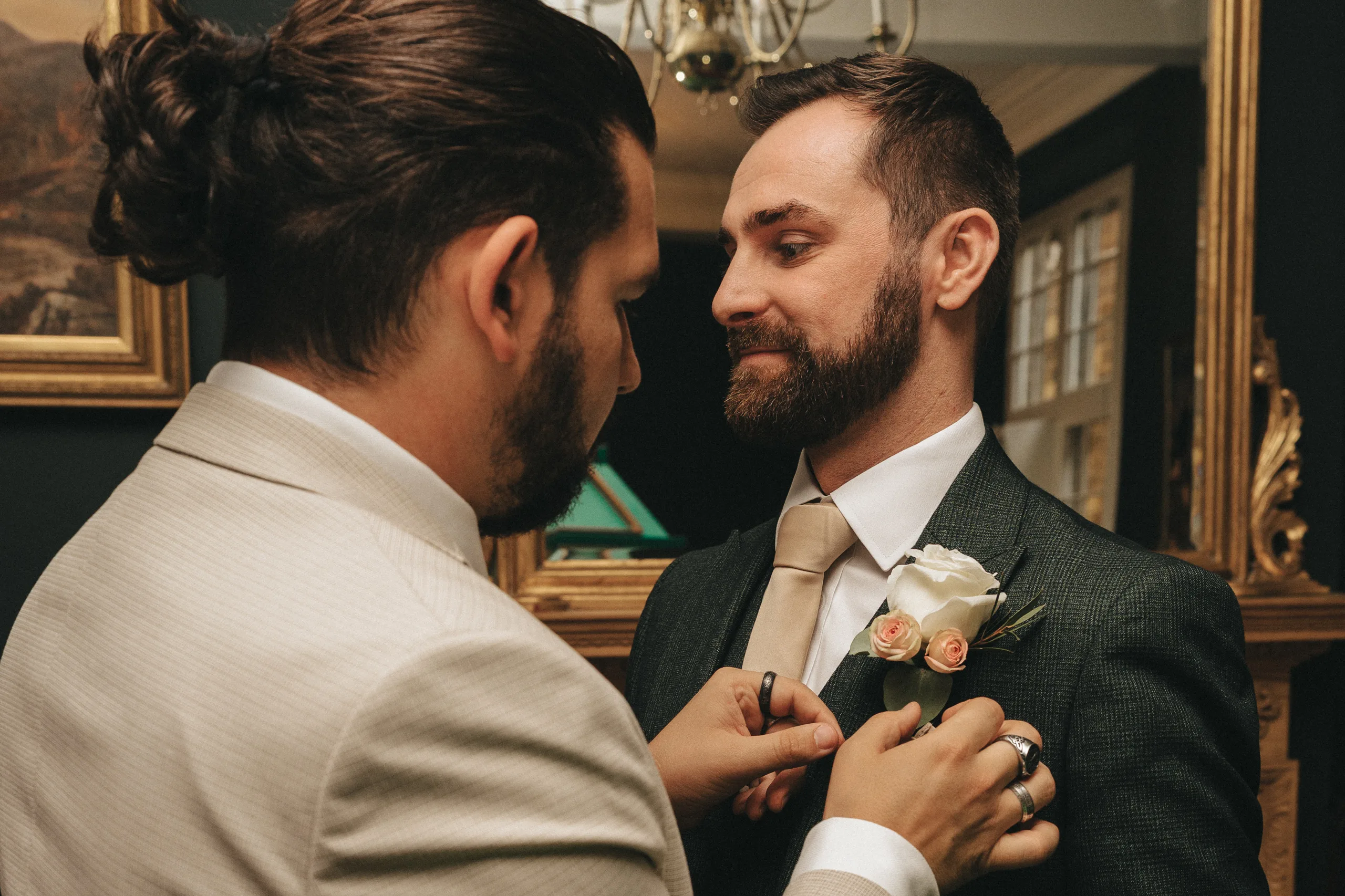 katie-adair-wedding-photography-rushton-hall-kettering-northamptonshire-30-of-1141-©-aimee-lince-photography-01-06-2025 Two men stand indoors as one, in a light suit, pins a boutonniere onto the other's dark green jacket. Captured in candid wedding photography, this elegant moment at a Rushton Hall wedding features gold-framed art in the background. © Aimee Lince Photography - Wedding photographer in Lincolnshire, Yorkshire & Nottinghamshire