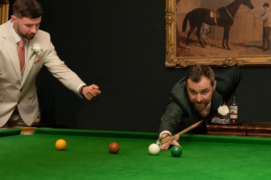 Two men dressed formally play snooker at a Rushton Hall wedding. One, in a cream suit with a boutonniere, gestures with his fist while the other lines up a shot. A painting of a horse and rider hangs on the wall behind them. © Aimee Lince Photography - Wedding photographer in Lincolnshire, Yorkshire & Nottinghamshire