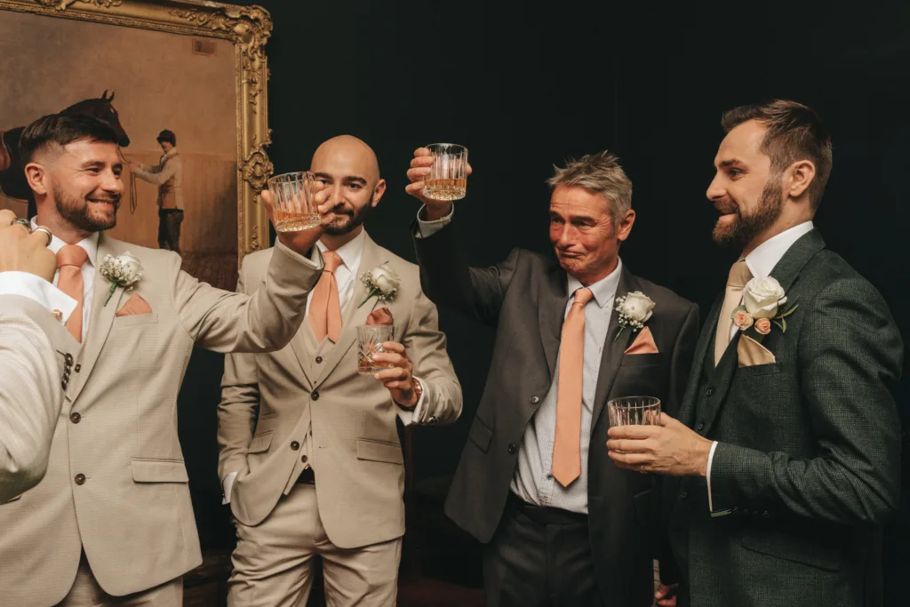 Four men in suits stand together indoors, smiling and raising glasses in a toast. Captured by a fun wedding photographer UK, three wear beige suits with peach ties, one wears a dark suit. A framed painting hangs on the dark wall behind them. © Aimee Lince Photography - Wedding photographer in Lincolnshire, Yorkshire & Nottinghamshire
