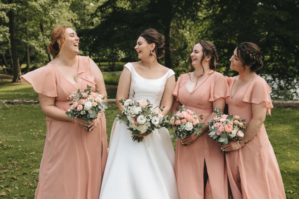 Four women stand outdoors on grass, smiling and holding bouquets. The woman in the center wears a white bridal gown; the three beside her wear matching peach dresses. Trees and sunlight fill the background, creating a cheerful, natural wedding scene. © Aimee Lince Photography - Wedding photographer in Lincolnshire, Yorkshire & Nottinghamshire