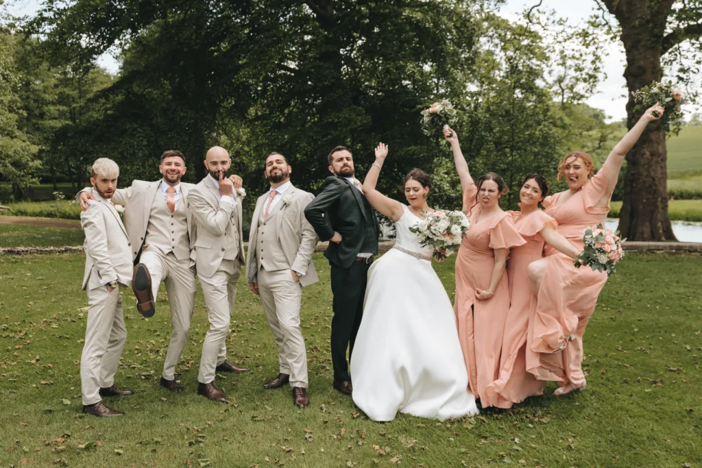 A wedding photographer in Kettering captures a playful outdoor moment—the groom in a dark green suit stands by the bride, surrounded by smiling groomsmen and bridesmaids who lift their arms or bouquets in celebration. © Aimee Lince Photography - Wedding photographer in Lincolnshire, Yorkshire & Nottinghamshire