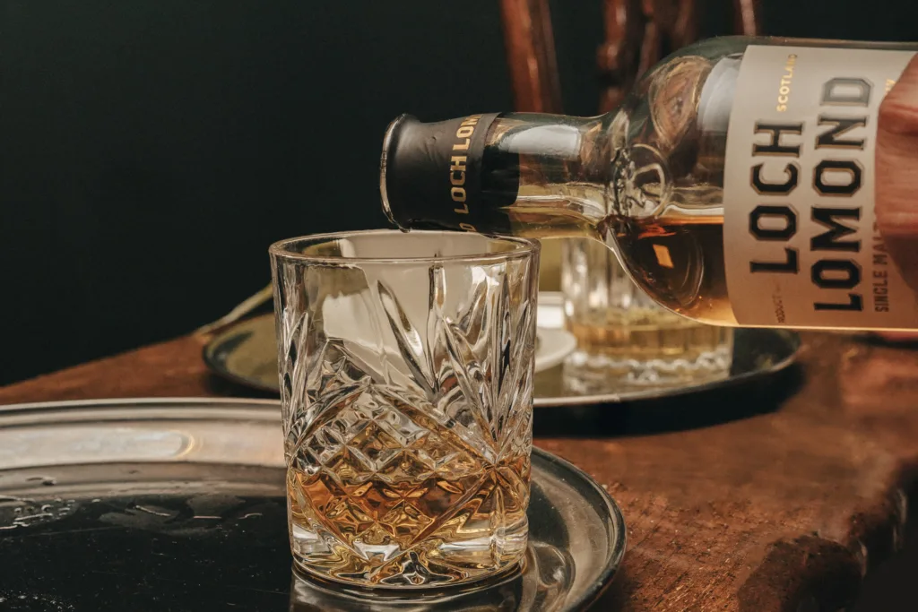 A hand pours Loch Lomond single malt Scotch whisky into a cut crystal glass, resting on a silver tray atop a wooden surface—much like the refined touches seen in candid wedding photography by a Kettering wedding photographer. © Aimee Lince Photography - Wedding photographer in Lincolnshire, Yorkshire & Nottinghamshire