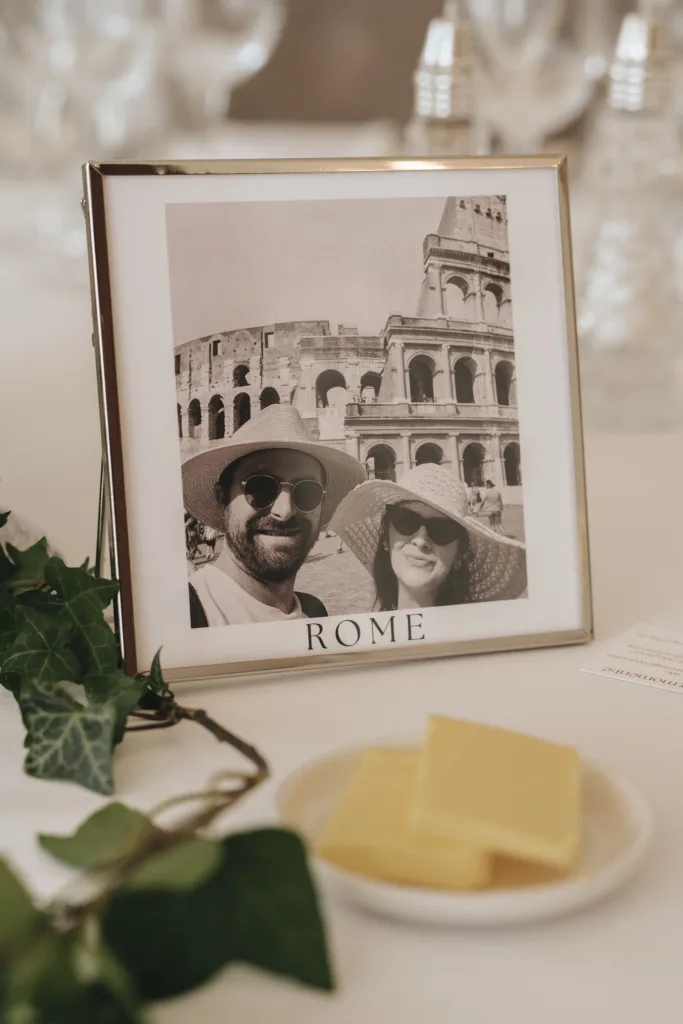 A framed black-and-white photo labeled “ROME” shows two people in sunglasses and hats smiling in front of the Colosseum. The frame sits on a table with a sprig of ivy, a note, and a small plate of rectangular butter pats in the foreground. © Aimee Lince Photography - Wedding photographer in Lincolnshire, Yorkshire & Nottinghamshire