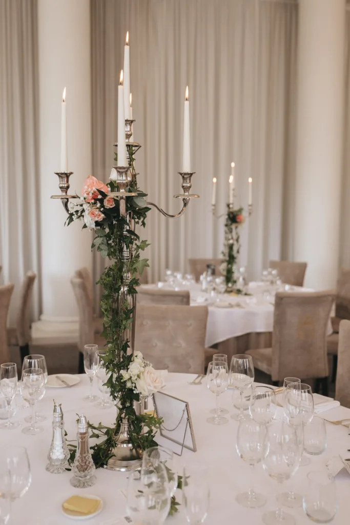 Elegant candelabra centerpiece with lit white candles, greenery, and pale pink flowers adorns a round table set with glassware and neutral-colored chairs in a softly lit, formal dining room with tall white columns and sheer curtains. © Aimee Lince Photography - Wedding photographer in Lincolnshire, Yorkshire & Nottinghamshire
