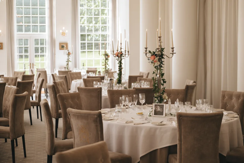 Elegant dining room with round tables covered in white cloths, set with glassware and cutlery. Velvet chairs surround each table. Tall candelabras with flowers serve as centerpieces. Large windows let in natural light, showcasing a lush green lawn outside. © Aimee Lince Photography - Wedding photographer in Lincolnshire, Yorkshire & Nottinghamshire