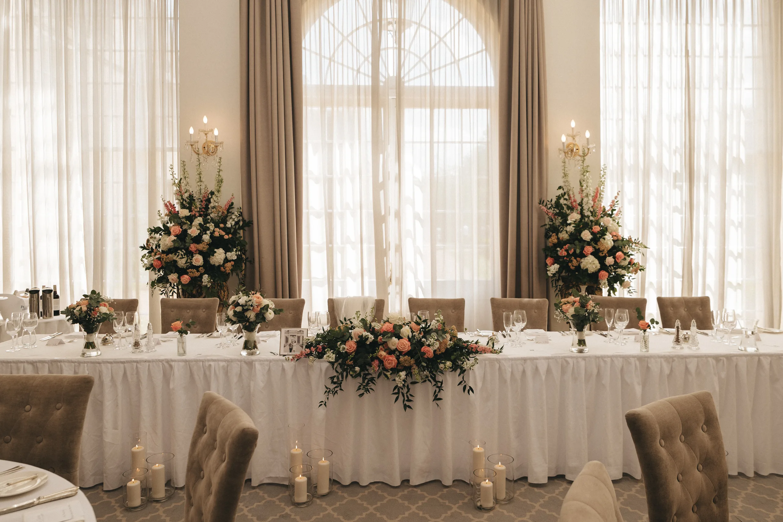 katie-adair-wedding-photography-rushton-hall-kettering-northamptonshire-497-of-1141-©-aimee-lince-photography-04-06-2025 A long, elegant banquet table at Rushton Hall is set with white cloth, pink and white flowers, glassware, and candles. Plush beige chairs surround the table as a photographer in Kettering captures the tall floral centerpieces by sheer curtained windows. © Aimee Lince Photography - Wedding photographer in Lincolnshire, Yorkshire & Nottinghamshire