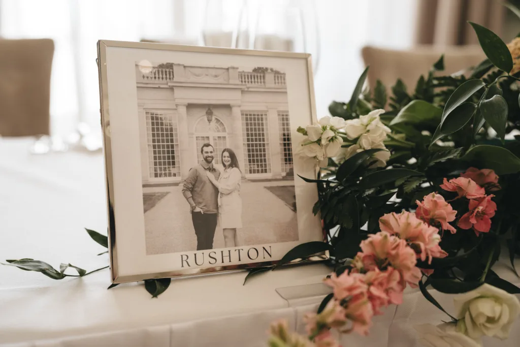 A framed black-and-white photo of a smiling couple stands on a table beside pink and white flowers and green leaves. The word “RUSHTON” is printed below the photo. The background is softly blurred, suggesting an elegant indoor setting. © Aimee Lince Photography - Wedding photographer in Lincolnshire, Yorkshire & Nottinghamshire