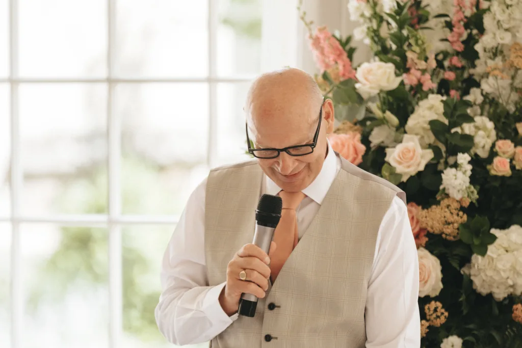 An older man in glasses, a light gray vest, and a peach tie holds a microphone and smiles while looking down. At a Rushton Hall wedding, he stands beside a large floral arrangement as sunlight pours through the window behind him. © Aimee Lince Photography - Wedding photographer in Lincolnshire, Yorkshire & Nottinghamshire