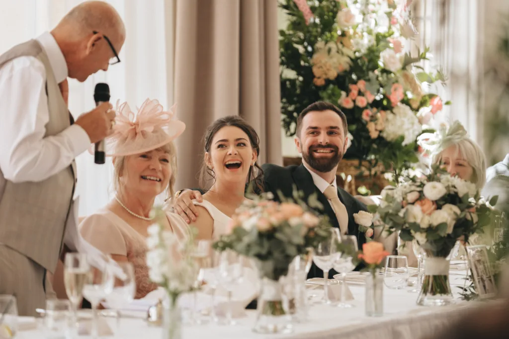 A wedding reception scene shows a smiling bride and groom seated at a table with family, listening to an older man giving a speech. The table is decorated with floral arrangements and glasses; a woman in a large pink hat sits beside the bride. Elegant flowers are in the background. © Aimee Lince Photography - Wedding photographer in Lincolnshire, Yorkshire & Nottinghamshire