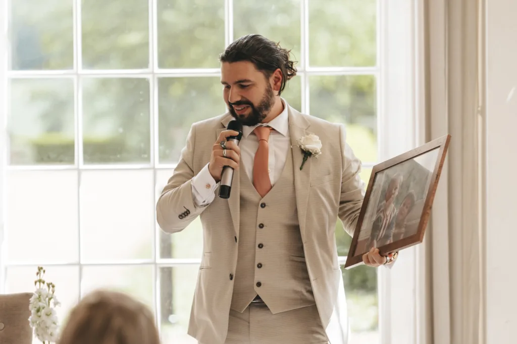 A man in a beige suit with a peach tie speaks into a microphone while holding a framed photo. By large window panels with greenery outside, he gives a heartfelt wedding speech—perfect for any photographer capturing special moments in Kettering. © Aimee Lince Photography - Wedding photographer in Lincolnshire, Yorkshire & Nottinghamshire