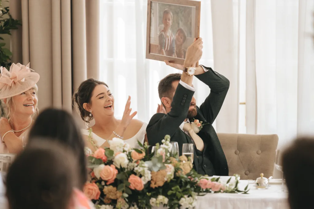 At a wedding reception in Kettering, a smiling bride and groom sit at a table with elegant floral arrangements. The groom holds up a framed childhood photo as the photographer captures the joyful bride and their guests in the foreground. © Aimee Lince Photography - Wedding photographer in Lincolnshire, Yorkshire & Nottinghamshire