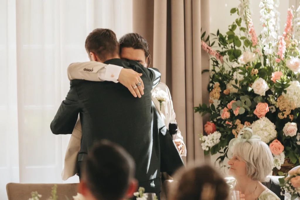 Two people wearing suits hug each other tightly at a Rushton Hall wedding near Kettering, standing beside a large floral arrangement. A woman with gray hair in white smiles warmly from a nearby table as soft sunlight fills the room, captured by the photographer. © Aimee Lince Photography - Wedding photographer in Lincolnshire, Yorkshire & Nottinghamshire