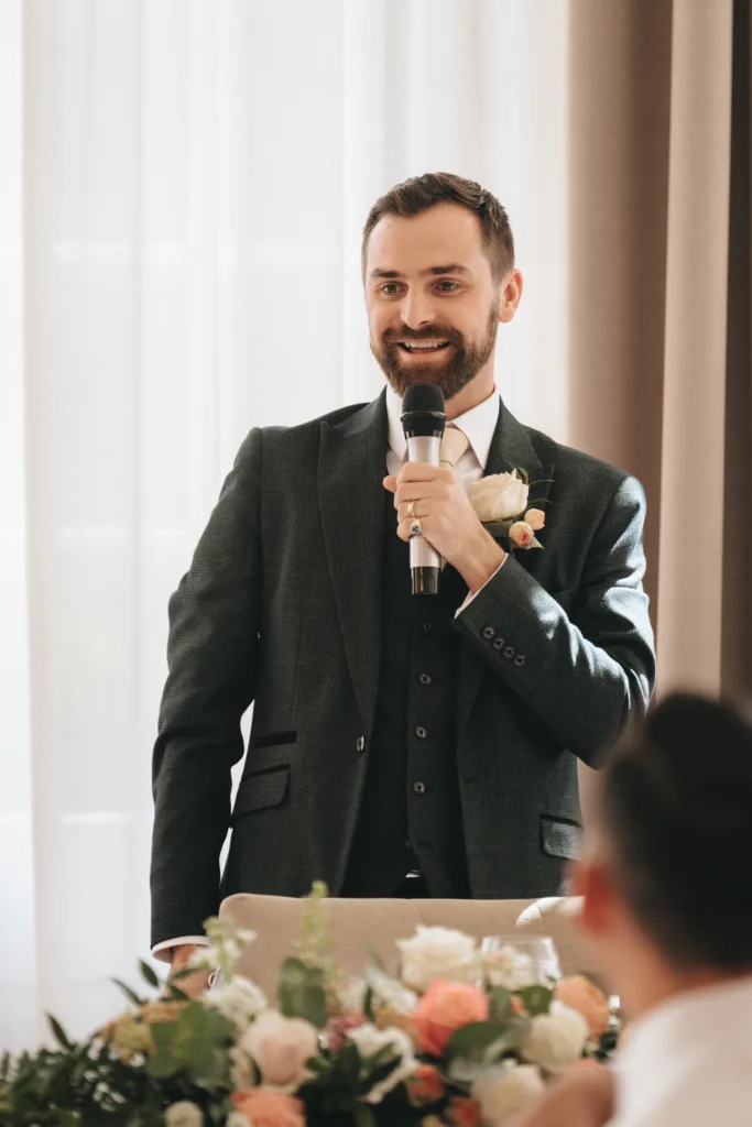 A bearded man in a dark suit and white shirt stands indoors at Rushton Hall, holding a microphone and smiling while giving a wedding speech. He wears a boutonnière near a table decorated with pastel flowers as light filters through sheer curtains behind him. © Aimee Lince Photography - Wedding photographer in Lincolnshire, Yorkshire & Nottinghamshire