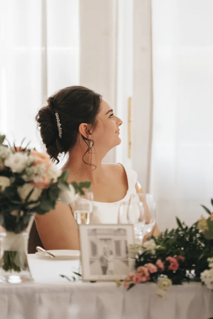 A bride with dark hair in an updo and a sparkling hairpiece smiles while sitting at a flower-adorned table at her Rushton Hall wedding. She wears a white off-the-shoulder dress; framed photos and glasses are visible on the table in front of her. © Aimee Lince Photography - Wedding photographer in Lincolnshire, Yorkshire & Nottinghamshire