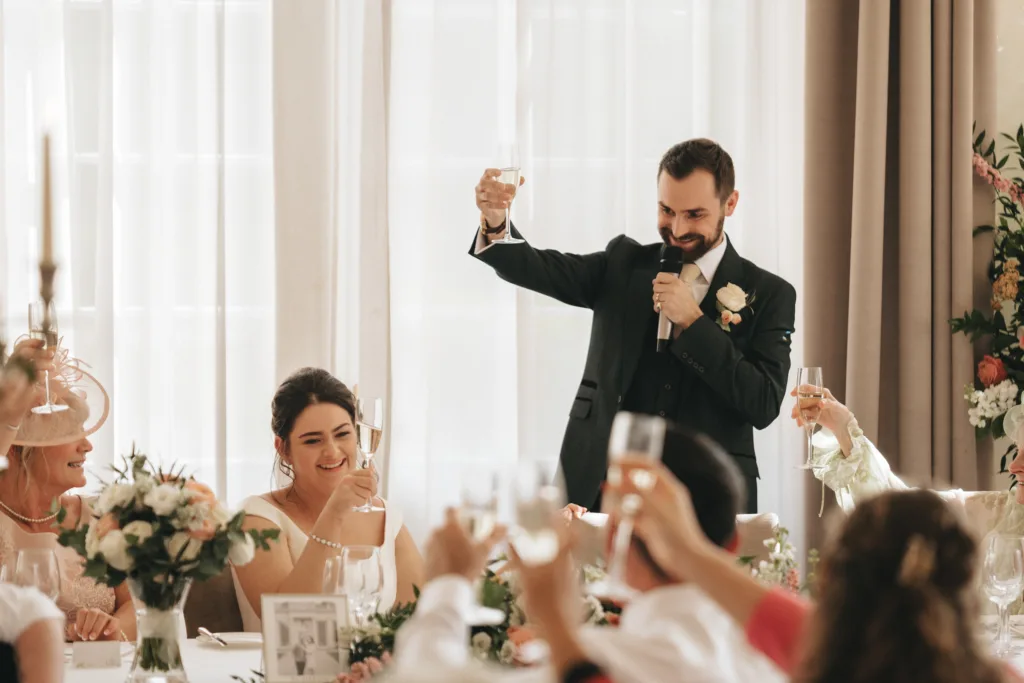 A man in a suit stands and gives a toast with a raised glass at a Rushton Hall wedding reception, smiling. Guests, including the bride and others, sit around the table and raise their glasses as the photographer captures the moment amid floral decor. © Aimee Lince Photography - Wedding photographer in Lincolnshire, Yorkshire & Nottinghamshire