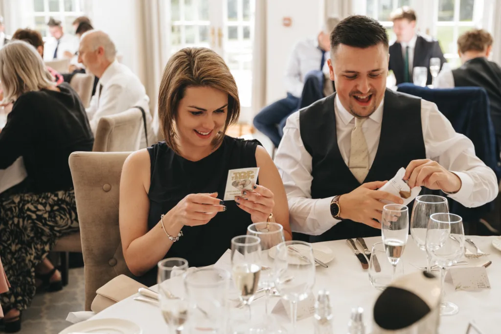 A woman and man dressed formally sit at a table set for a meal, smiling while opening small gifts. The table has wine glasses, cutlery, and elegant decor. Other guests are visible in the bright, airy background, suggesting a celebratory event. © Aimee Lince Photography - Wedding photographer in Lincolnshire, Yorkshire & Nottinghamshire