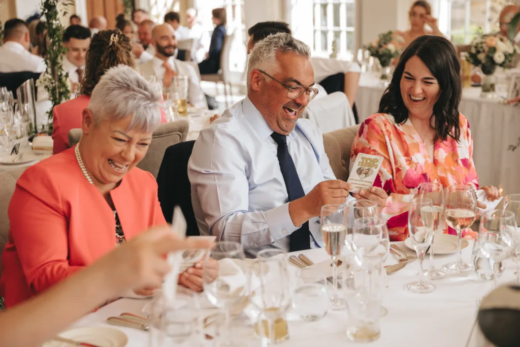 Three adults in formal attire sit at a banquet table, laughing and smiling as two of them hold scratch-off lottery tickets. The table is set with many glasses and plates, and other guests can be seen enjoying the festive atmosphere in the background. © Aimee Lince Photography - Wedding photographer in Lincolnshire, Yorkshire & Nottinghamshire