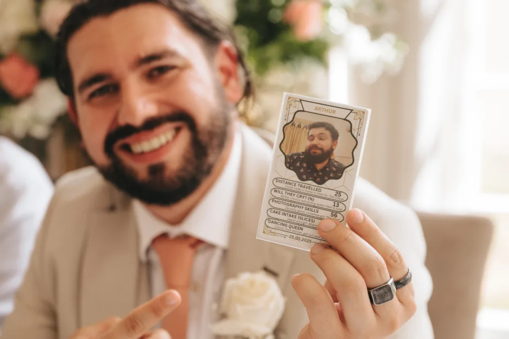 A smiling man in a light suit and peach tie points at a custom playing card with his photo and the name “Arthur.” The card features stats and text below the photo. The man sits indoors, wearing a white rose boutonniere, with a floral background. © Aimee Lince Photography - Wedding photographer in Lincolnshire, Yorkshire & Nottinghamshire