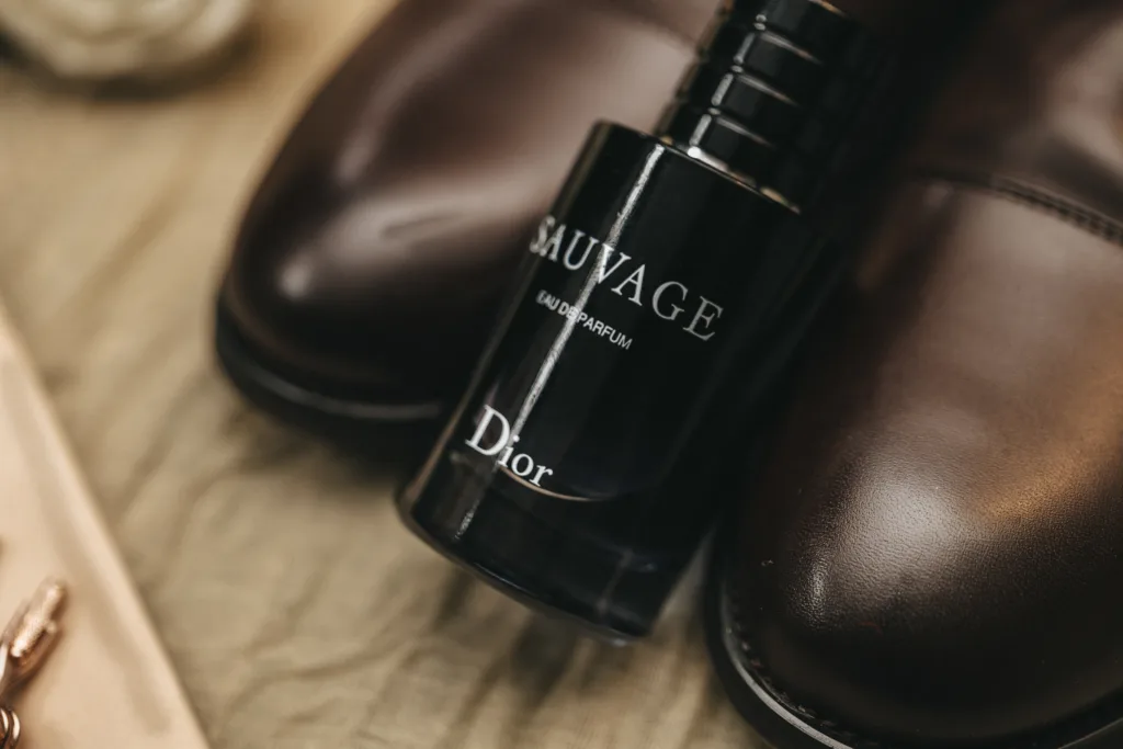 A black bottle of Dior Sauvage Eau de Parfum stands upright between polished brown leather dress shoes on a beige fabric surface—an artful composition that could inspire any candid wedding photography for a fun wedding photographer UK. © Aimee Lince Photography - Wedding photographer in Lincolnshire, Yorkshire & Nottinghamshire