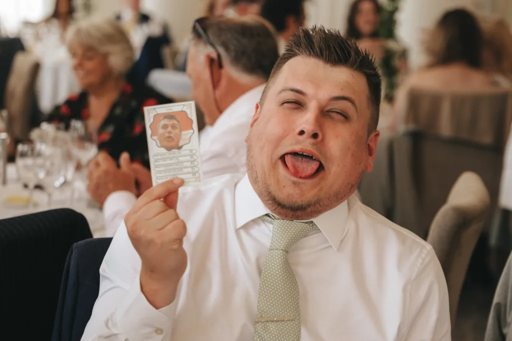 A man in a white shirt and light green tie sits at a table, making a silly face with his tongue out. He holds up a card featuring a photo of a face with a similar expression. People sit and talk in the blurred background at what appears to be a social event. © Aimee Lince Photography - Wedding photographer in Lincolnshire, Yorkshire & Nottinghamshire