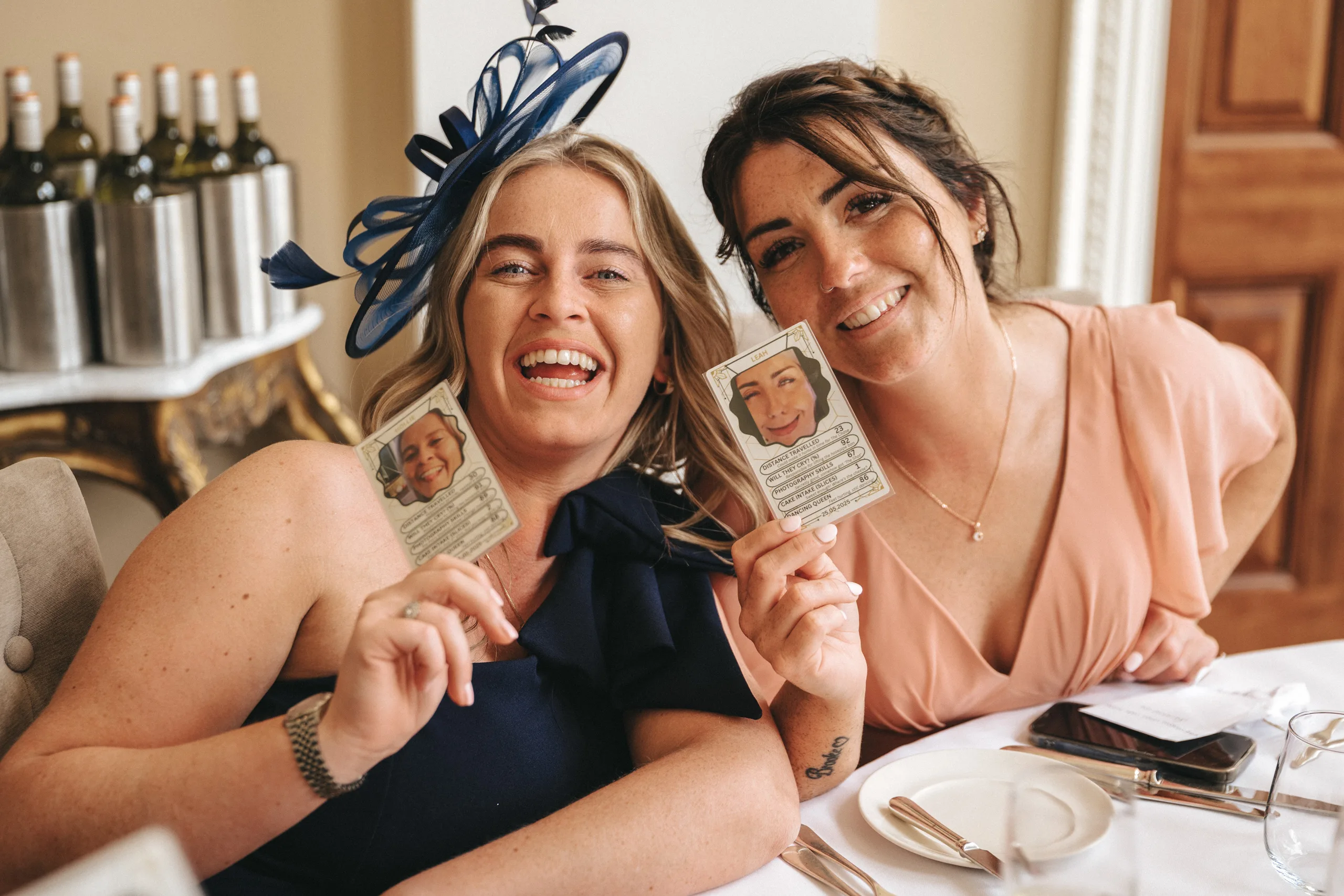 katie-adair-wedding-photography-rushton-hall-kettering-northamptonshire-606-of-1141-©-aimee-lince-photography-04-06-2025 Two women sitting at a table, smiling and holding up cards with cartoon-like faces and text. One woman wears a navy dress with a blue fascinator, the other a peach dress. Wine bottles and cutlery are visible in the elegant indoor setting. © Aimee Lince Photography - Wedding photographer in Lincolnshire, Yorkshire & Nottinghamshire