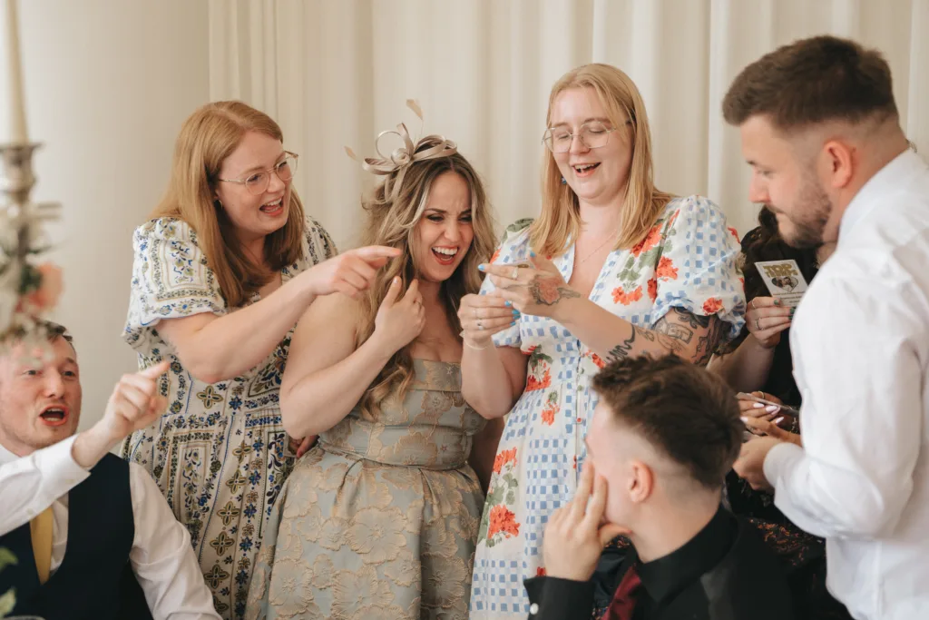 A group of people at a celebration laugh and point while gathered closely together. Two women in floral dresses and one in a beige patterned dress are central, reacting joyfully. Two men, one seated and one standing, also laugh. The atmosphere is cheerful and lively. © Aimee Lince Photography - Wedding photographer in Lincolnshire, Yorkshire & Nottinghamshire