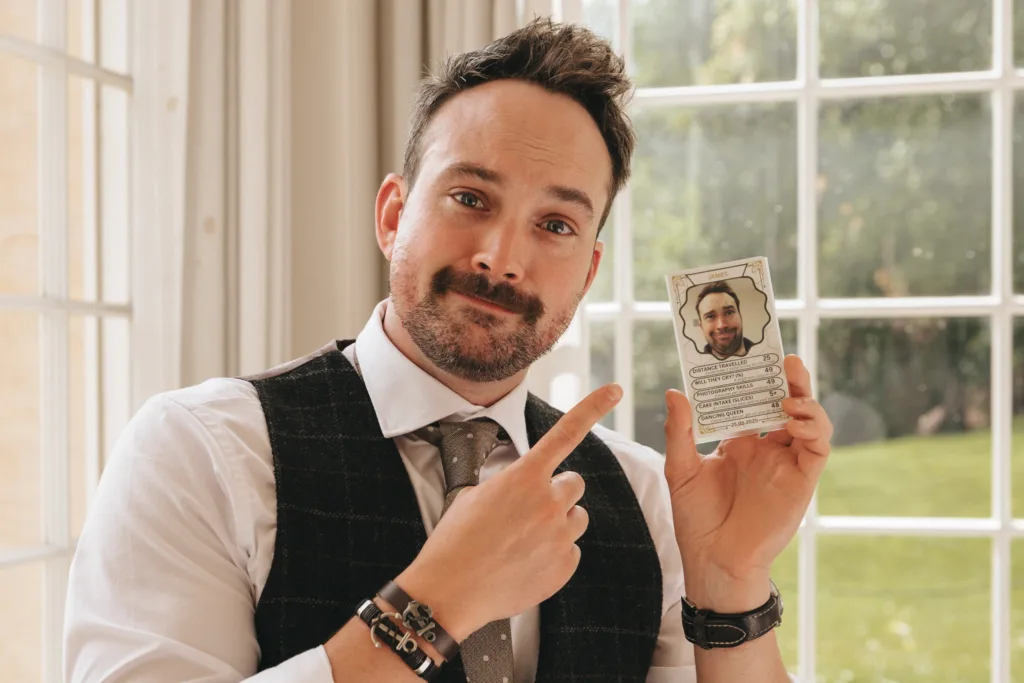A man in a dress shirt, vest, and tie stands indoors by large windows, smiling and pointing at a card he holds up with his left hand. The card features his photo and some text. Bright daylight and green grass are visible through the windows. © Aimee Lince Photography - Wedding photographer in Lincolnshire, Yorkshire & Nottinghamshire