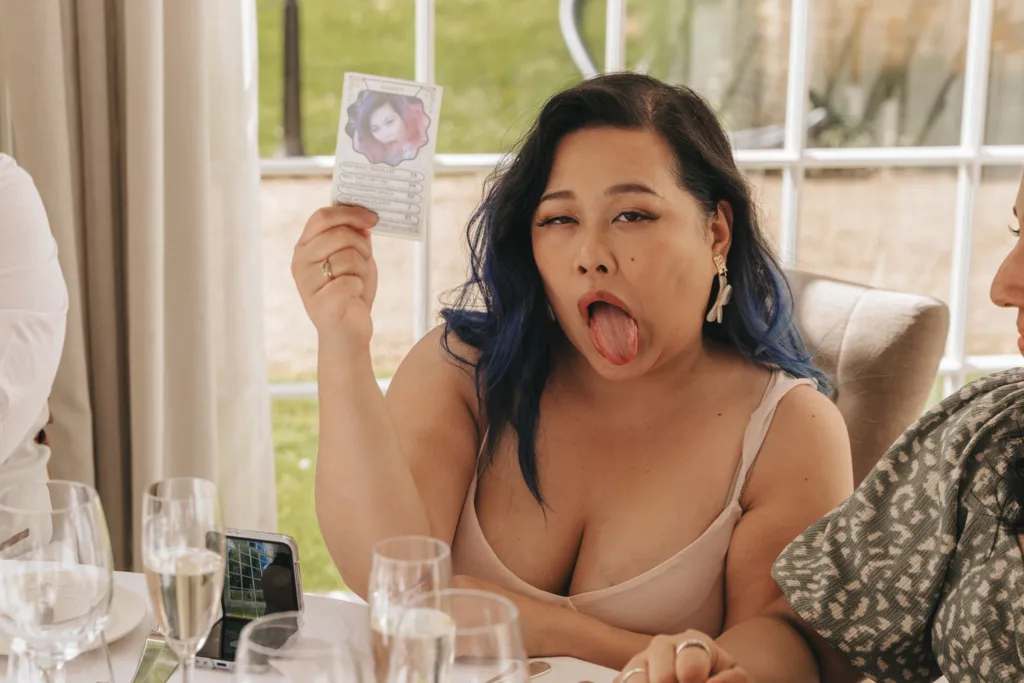 A woman with long dark hair and blue tips sits at a dining table, sticking out her tongue playfully and holding up a card with her photo on it. She wears a cream-colored top and unique earrings. Table settings and glasses are visible in front of her. © Aimee Lince Photography - Wedding photographer in Lincolnshire, Yorkshire & Nottinghamshire