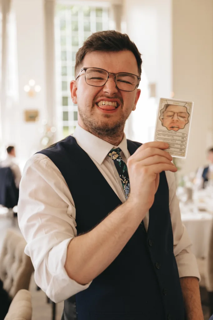 A man in glasses and a navy vest smiles playfully, showing his teeth, while holding up a card with a cartoon face in front of his own. The background shows a bright, elegant room with large windows, white walls, and tables set for an event. © Aimee Lince Photography - Wedding photographer in Lincolnshire, Yorkshire & Nottinghamshire