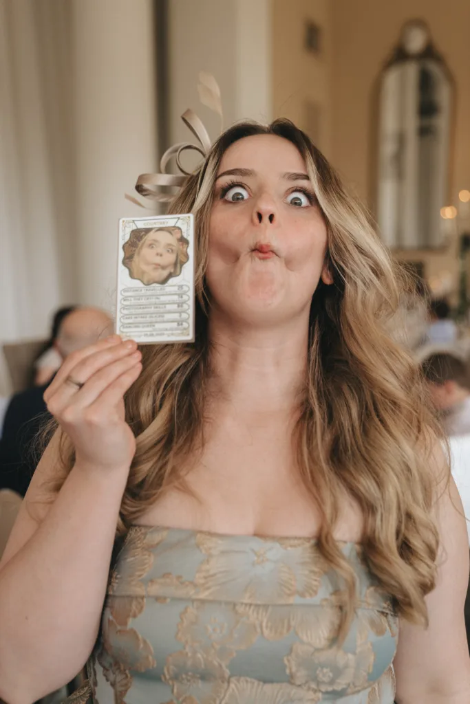 A woman with long wavy blonde hair wearing a strapless patterned dress and fascinator holds a card with a funny face on it in front of her chest. She mimics the card’s facial expression, puffing out her cheeks and widening her eyes in a playful manner. © Aimee Lince Photography - Wedding photographer in Lincolnshire, Yorkshire & Nottinghamshire