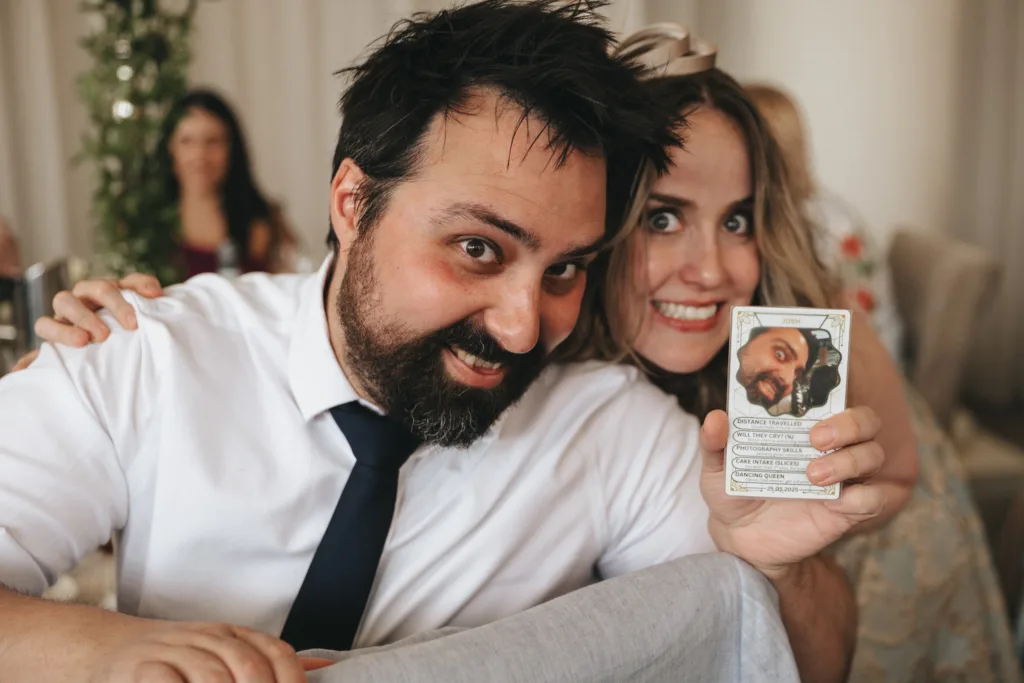 A smiling man with dark hair and a beard sits beside a smiling woman with long hair. The woman holds up a card featuring the man’s face and text. Both wear dress clothes and appear happy. Other people and greenery are visible in the softly focused background. © Aimee Lince Photography - Wedding photographer in Lincolnshire, Yorkshire & Nottinghamshire