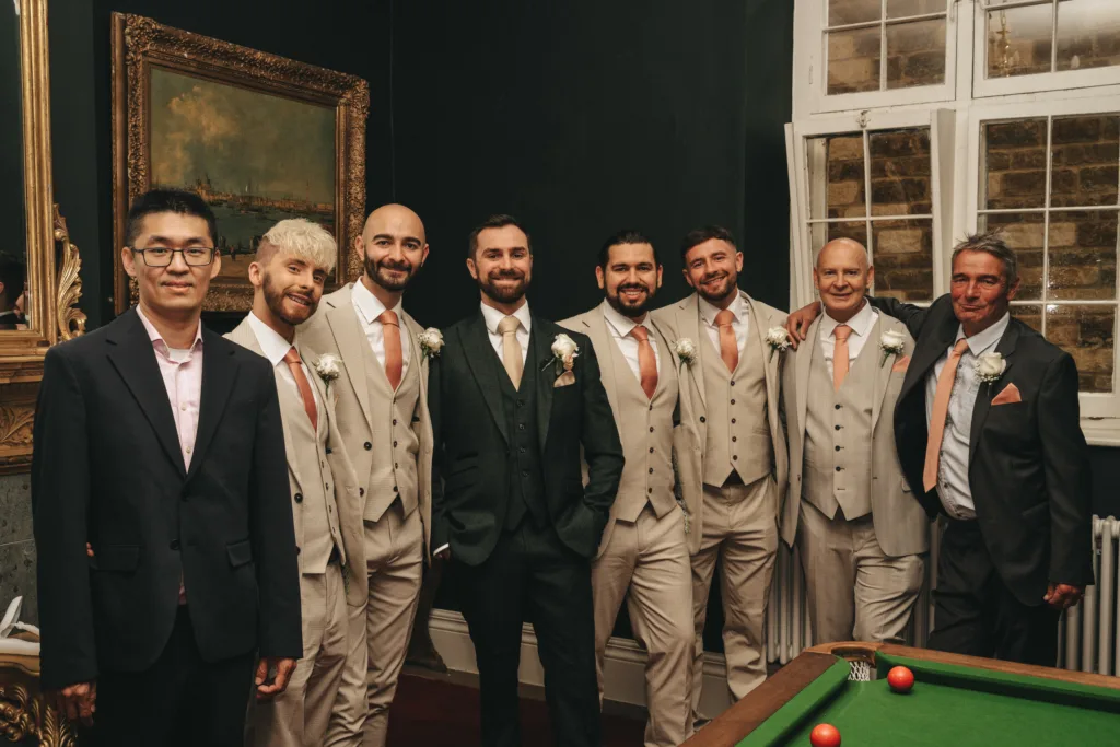 Seven men pose together in a dimly lit room near a pool table at a Rushton Hall wedding. Six wear matching beige suits with peach ties; one in the center wears dark green. Captured by a Kettering wedding photographer, their candid smiles shine. © Aimee Lince Photography - Wedding photographer in Lincolnshire, Yorkshire & Nottinghamshire