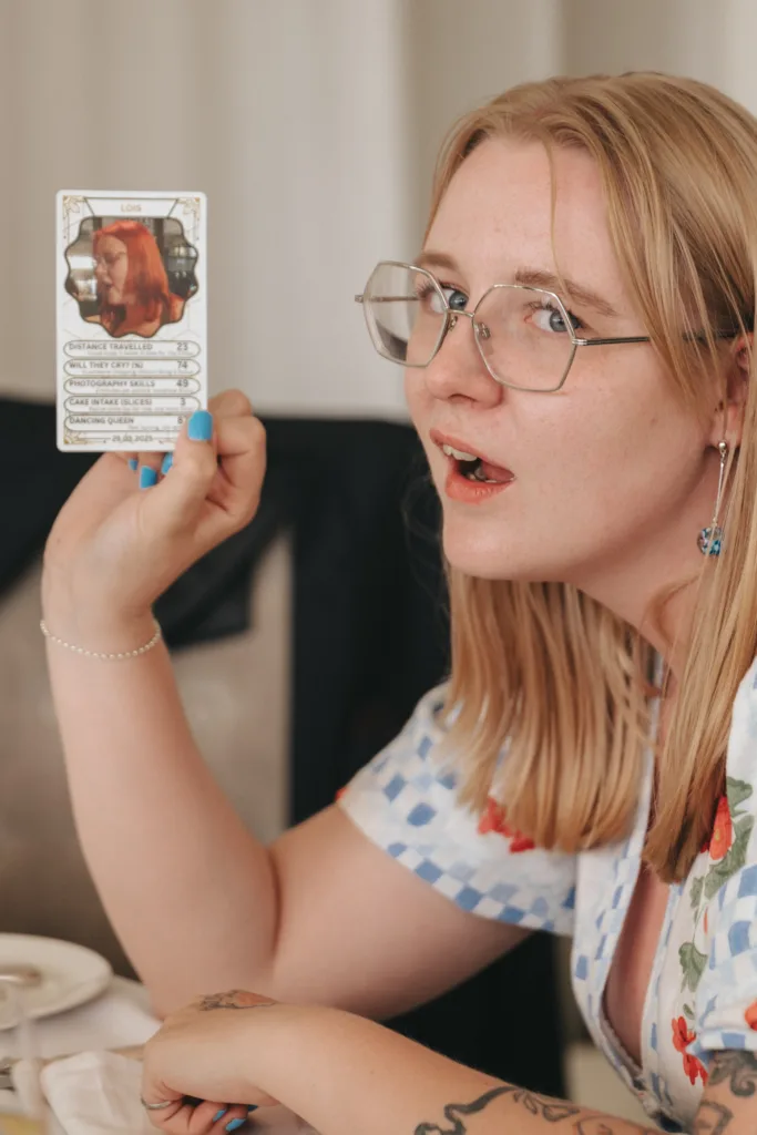 A blonde woman with glasses and floral clothing holds up a card with a red-haired woman's portrait and stats. She looks engaged, mouth open slightly, and is sitting at a table with white dishes, her nails painted blue, and tattoos visible on her arm. © Aimee Lince Photography - Wedding photographer in Lincolnshire, Yorkshire & Nottinghamshire