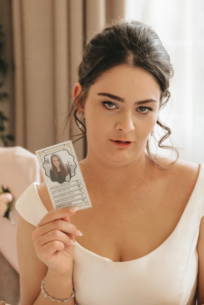 A woman in a white dress with dark hair styled up makes a humorous face while holding a card with a photo and text on it. She is indoors, in soft natural light, with beige and white decor visible in the background. © Aimee Lince Photography - Wedding photographer in Lincolnshire, Yorkshire & Nottinghamshire