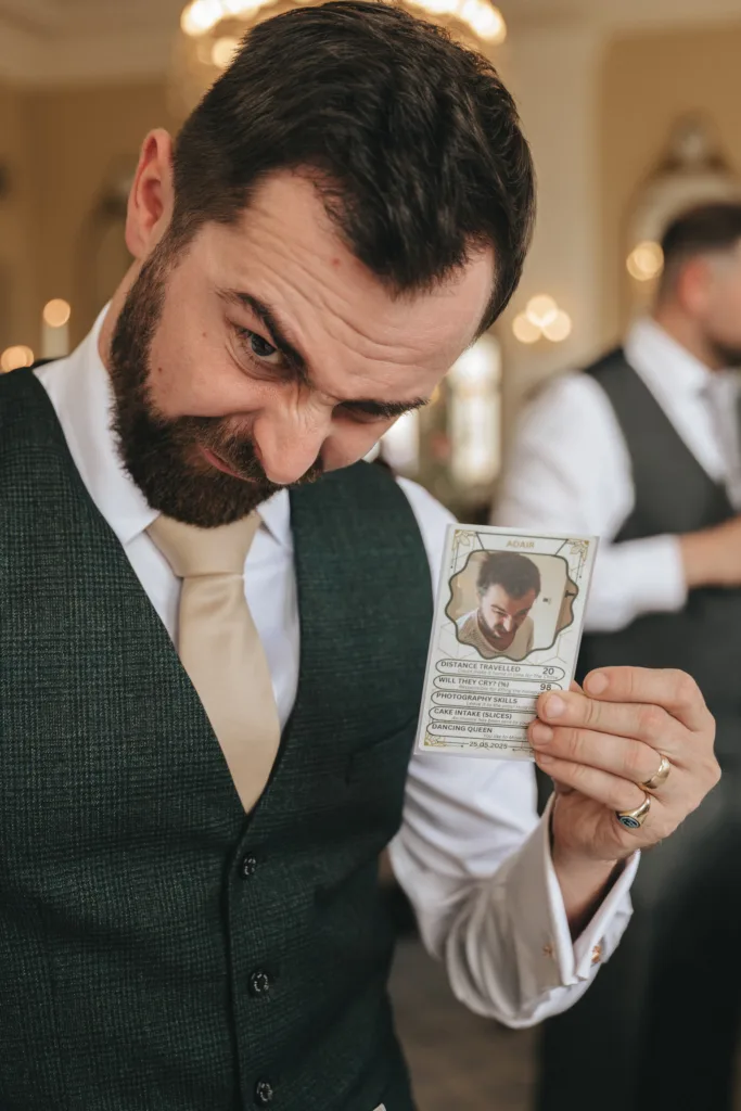 A bearded man in a dark vest, white shirt, and beige tie holds up a custom card featuring his portrait and stats. He raises one eyebrow and smirks, tilting his head playfully. The background is softly lit, with another person blurred behind him. © Aimee Lince Photography - Wedding photographer in Lincolnshire, Yorkshire & Nottinghamshire
