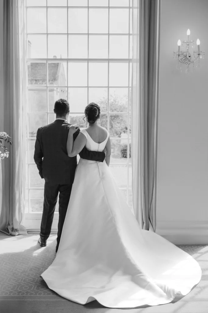 A bride in a flowing white gown stands beside her groom at Rushton Hall, both facing a large window with light streaming in. The photographer captures her hand on his shoulder, elegant curtains framing the scene and a chandelier hanging to the right. © Aimee Lince Photography - Wedding photographer in Lincolnshire, Yorkshire & Nottinghamshire