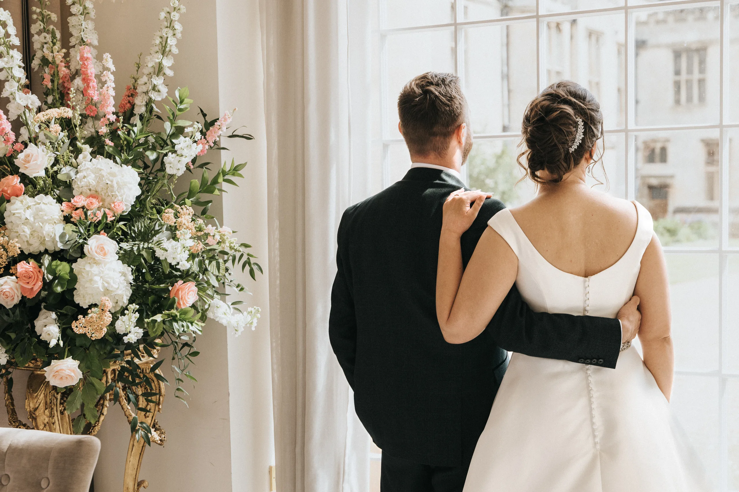 katie-adair-wedding-photography-rushton-hall-kettering-northamptonshire-648-of-1141-©-aimee-lince-photography-05-06-2025 A bride in a white dress and groom in a dark suit stand with their backs to the camera, gazing out a large window. Captured by a wedding photographer, the scene features a large arrangement of white, pink, and peach flowers to the left. © Aimee Lince Photography - Wedding photographer in Lincolnshire, Yorkshire & Nottinghamshire
