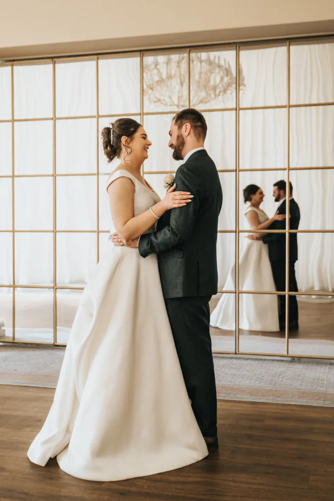 A bride in a white gown and a groom in a dark suit stand smiling and holding each other in front of a large, gold-framed mirror wall at Rushton Hall, Kettering. Their reflections and a faint chandelier add elegance to their wedding moment. © Aimee Lince Photography - Wedding photographer in Lincolnshire, Yorkshire & Nottinghamshire