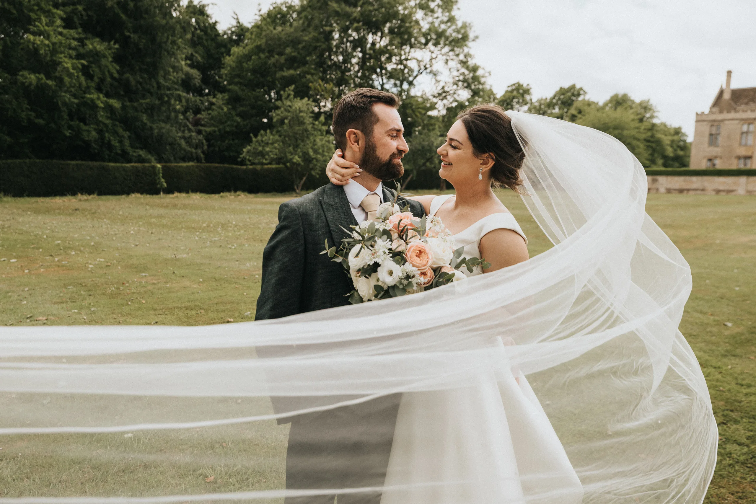katie-adair-wedding-photography-rushton-hall-kettering-northamptonshire-677-of-1141-©-aimee-lince-photography-05-06-2025 A bride and groom stand outdoors on grass at Rushton Hall, gazing lovingly at each other. The bride’s veil billows dramatically as a Kettering wedding photographer captures the moment with historic buildings and trees in the background. © Aimee Lince Photography - Wedding photographer in Lincolnshire, Yorkshire & Nottinghamshire