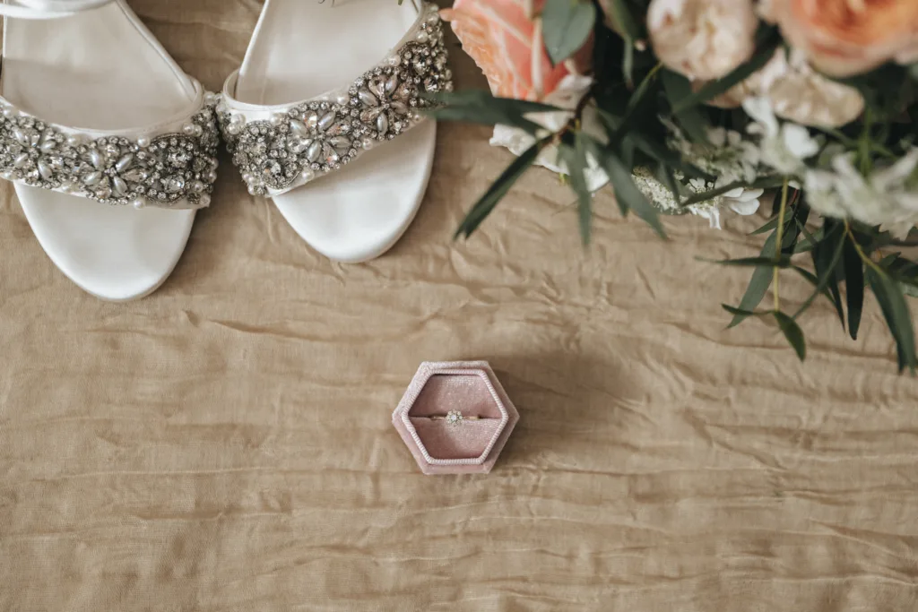 White sandals with jeweled straps and a bouquet of flowers rest on beige cloth. A pink hexagonal ring box with a diamond ring sits at the center, perfect for an east midlands wedding photographer to capture romantic moments. © Aimee Lince Photography - Wedding photographer in Lincolnshire, Yorkshire & Nottinghamshire