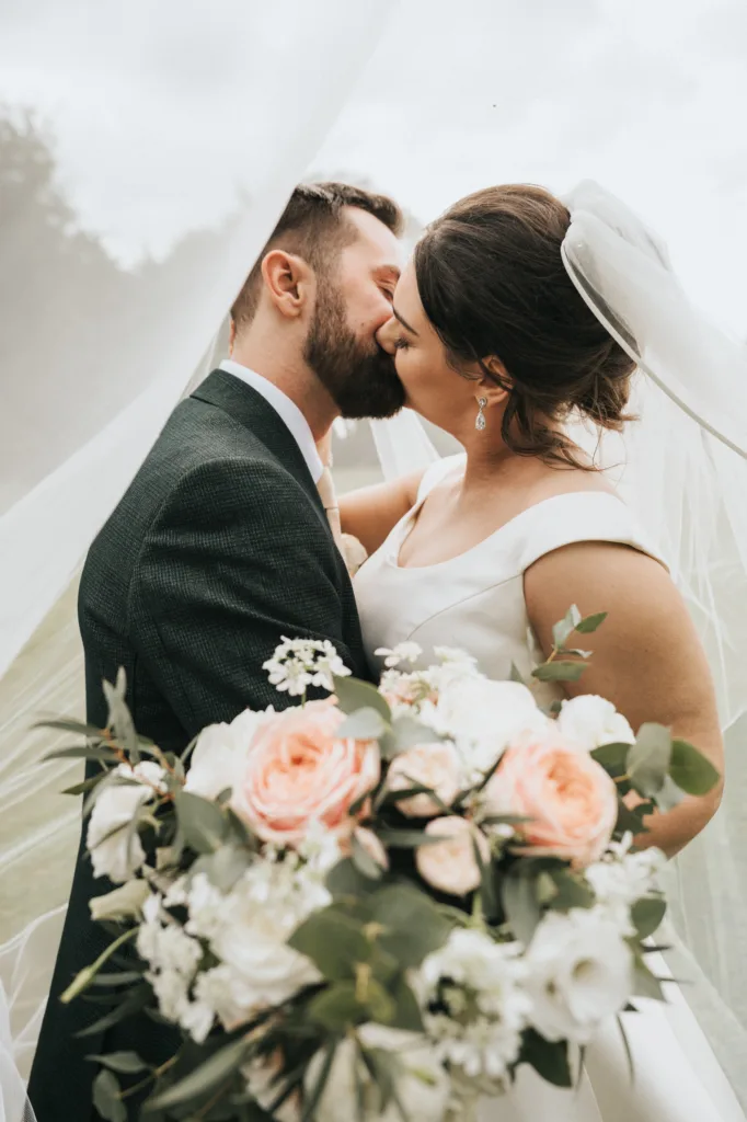 A bride and groom share a kiss under a white veil outdoors at Rushton Hall. The wedding photographer captures the bride in a white dress holding pink and white flowers, while soft, natural light creates a romantic atmosphere. © Aimee Lince Photography - Wedding photographer in Lincolnshire, Yorkshire & Nottinghamshire