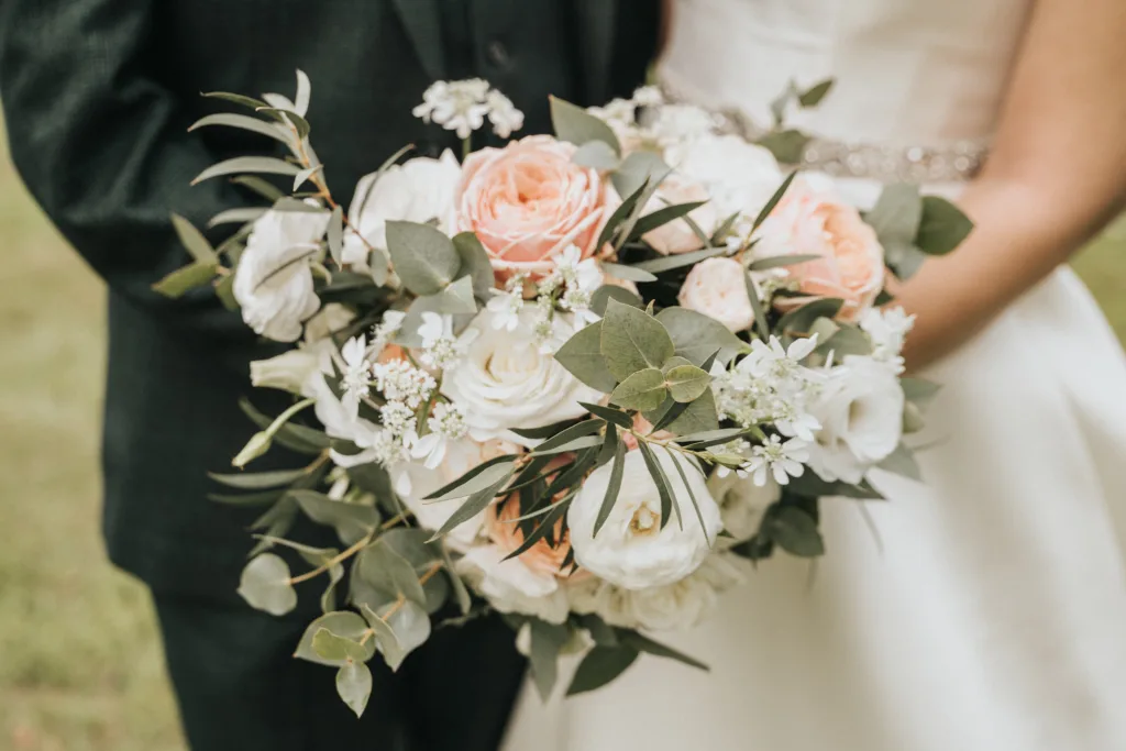 A bride in a white dress holds a bouquet of pale pink and white roses, white ranunculus, small white flowers, and green foliage. A wedding photographer in Kettering captures only the upper torsos of the bride and a guest in a dark suit. © Aimee Lince Photography - Wedding photographer in Lincolnshire, Yorkshire & Nottinghamshire